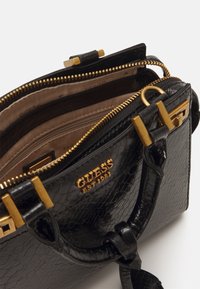 Musta käsilaukku, jossa on matelijannahkatekstuuri, kultaiset metalliosat, vetoketjukiinnitys sekä ruskea sisus, jossa on osastot ja brändilogo.