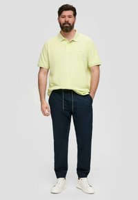 Jasnożółta koszulka polo z subtelnym logo, zestawiona z granatowymi spodniami i białymi trampkami. Tkanina wydaje się miękka i ma luźny krój.