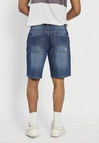 Bărbat purtând pantaloni scurți din denim albastru, șosete albe și adidași albi Nike, stând cu spatele la un fundal alb.