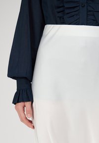 Bluză bleumarin cu față volanată și mâneci bufante; combinată cu o fustă netedă, mătăsoasă, de culoare deschisă. Prim-plan al unei mâini pe țesătură.