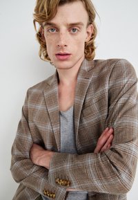 Brown plaid blazer with a textured fabric. Features blue and white stripes, gold button accents, and a gray t-shirt underneath. Crossed arms.