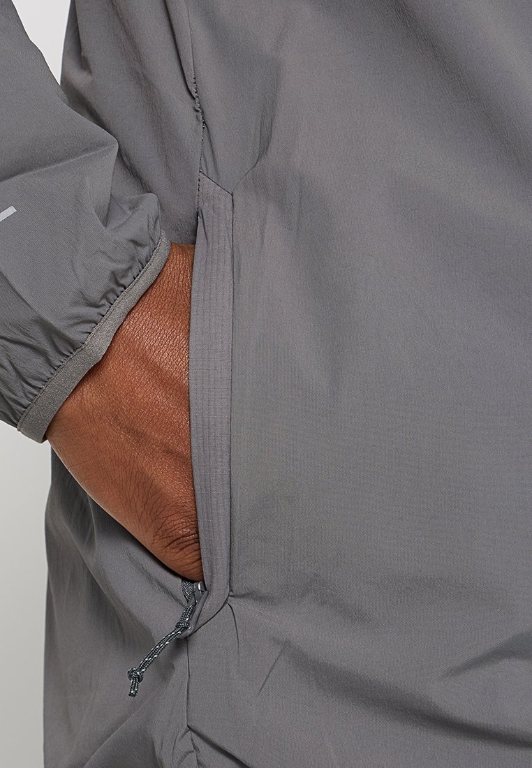 Graue, leichte Jacke mit elastischen Bündchen und einer Seitentasche mit Kordelzug. Glattes Material, minimalistisches Design und praktische Details.