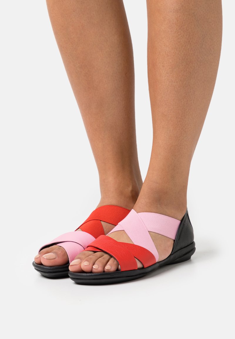 Röda och rosa elastiska remmar korsar en svart sula sandal, vilket visar en slät yta och en öppen tå-design.