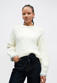 Kvinne med flette hår iført hvit genser og mørke jeans med et svart stoffblomsterbelte, poserer med en hånd i lommen.