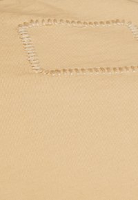 Beige tyg med en ribbad textur har en rektangulär broderad detalj. Stygnens färg är ljusare, vilket skapar en subtil kontrast.