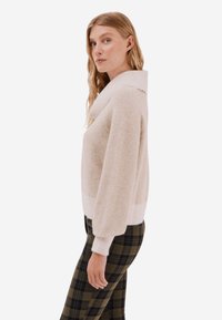 Beżowy sweter dzianinowy z szerokim kołnierzem i ściągaczami, o miękkiej teksturze. W połączeniu z kraciastymi spodniami w ciemnozielonym i czarnym kolorze.