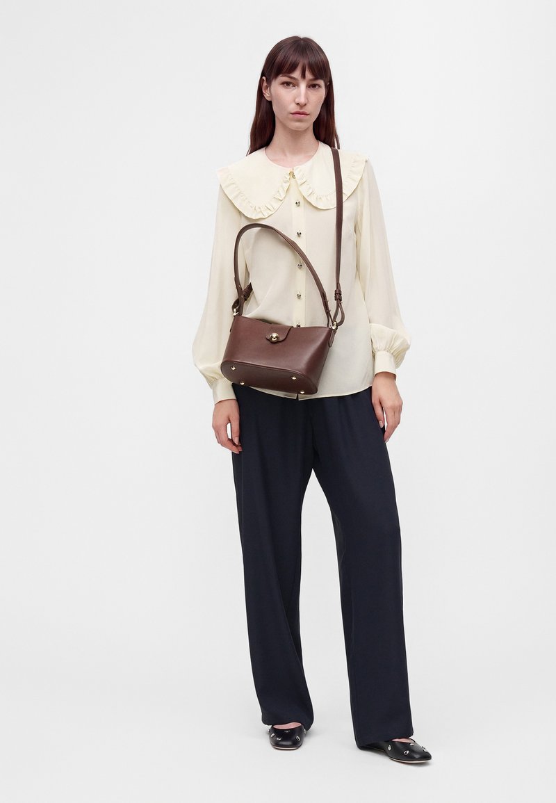 Femme portant une blouse crème avec un grand col, un pantalon noir à jambes larges, des chaussures noires, et portant un sac bandoulière en cuir marron avec un fermoir doré.