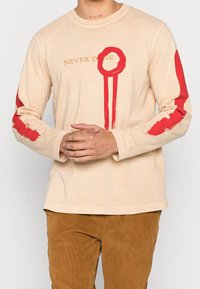 Beige pitkähihaisessa t-paidassa on punaisia maalin kaltaisia yksityiskohtia hihoissa, ja siinä on teksti "NEVER DONE" kullassa. Pehmeä ja rento kangas.