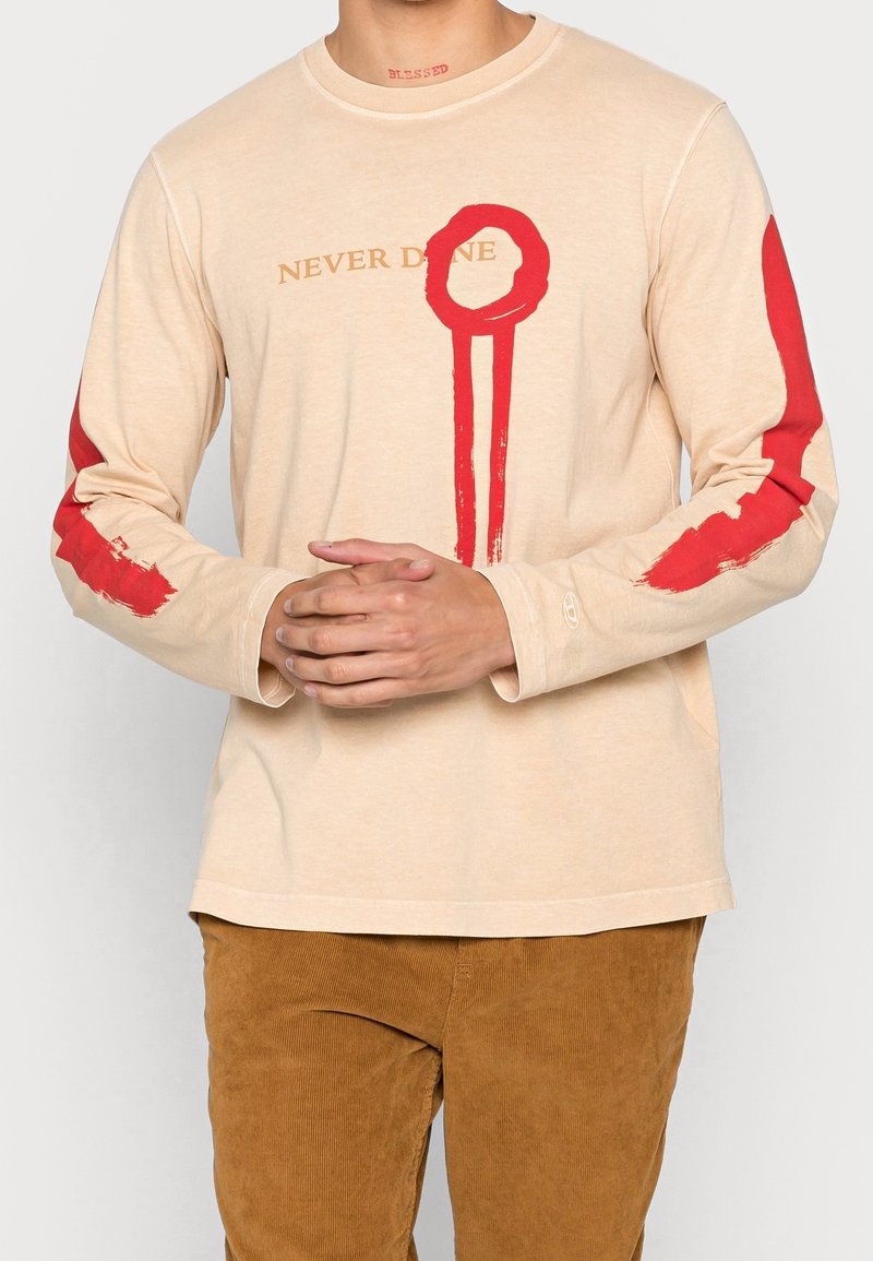Beige pitkähihaisessa t-paidassa on punaisia maalin kaltaisia yksityiskohtia hihoissa, ja siinä on teksti "NEVER DONE" kullassa. Pehmeä ja rento kangas.