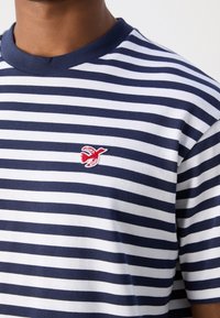 Marineblå og hvid stribet T-shirt med rund halsudskæring, korte ærmer og et lille rødt fuglelogo broderet på brystet.