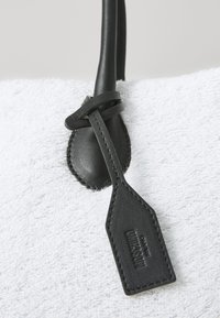 Čierna kožená visačka s reťazovým detailom, pripevnená na textúrovanej bielej tkanine, s prešitým okrajom a embosovanou značkou.