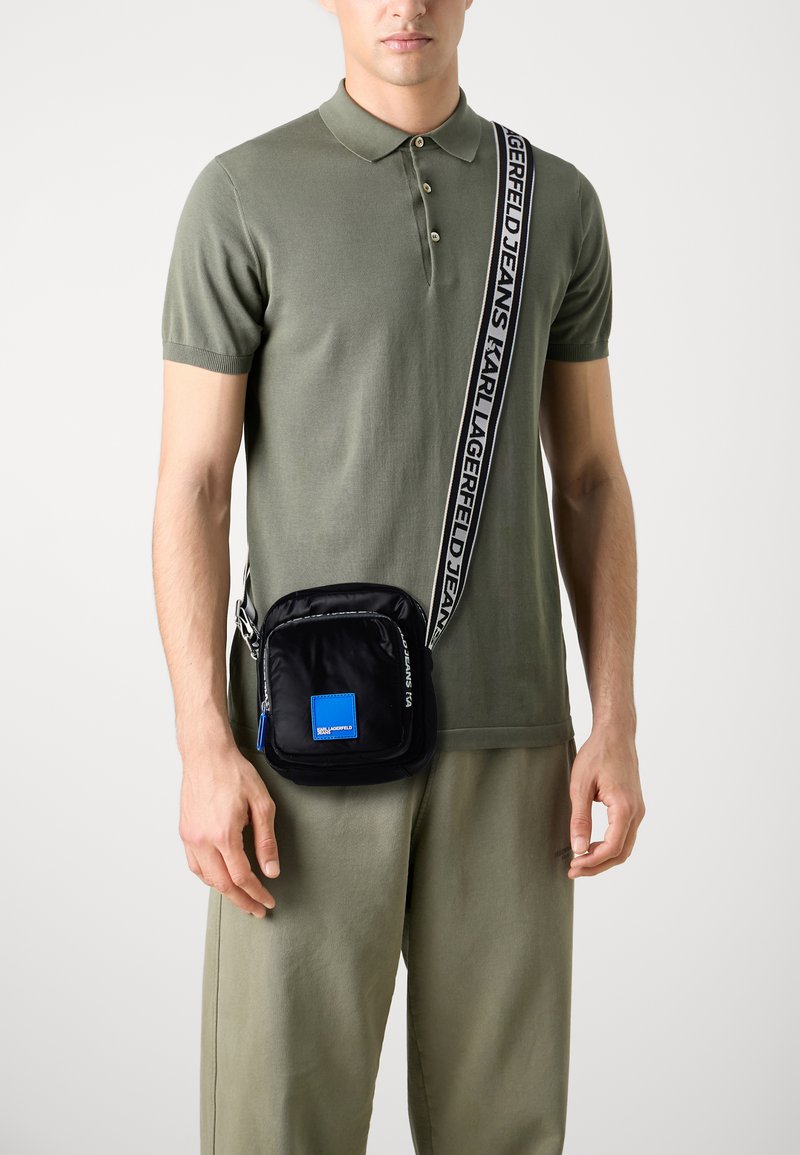 Črna najlon torba čez ramo z modrim logotipnim nalepkom, nastavljiv pas z napisom "KARL LAGERFELD JEANS" in zadrgo.