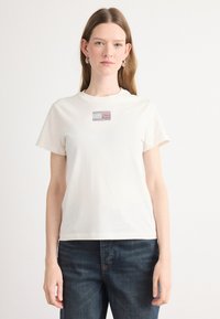Kortærmet t-shirt med rund hals i off-white bomuld med et lille, farverigt rhinsten-design på fronten, parret med mørkeblå jeans.