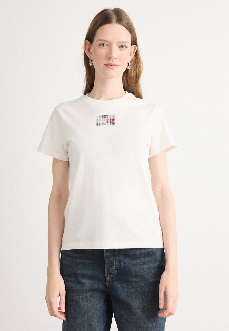 Kortærmet t-shirt med rund hals i off-white bomuld med et lille, farverigt rhinsten-design på fronten, parret med mørkeblå jeans.