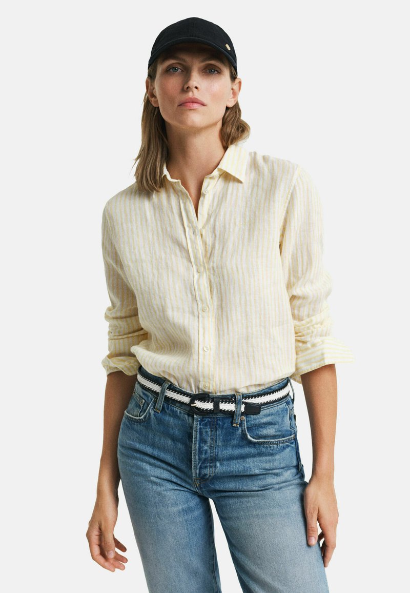 Gestreepte gele button-down shirt met opgerolde mouwen, gecombineerd met lichtblauwe jeans en een zwarte pet. Bevat een geweven riem met accenten.