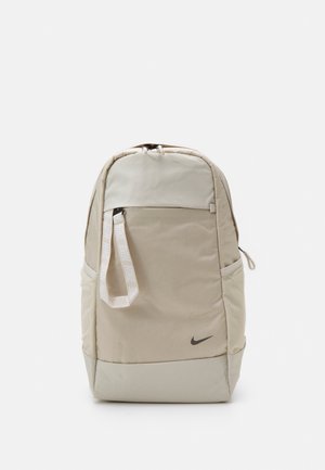 Beżowy plecak Nike z przednią kieszenią, białym paskiem z logo, bocznymi kieszeniami oraz czarnym logo Nike w kształcie łyżwy w dolnym prawym rogu.