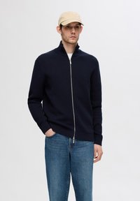 Maglione navy con zip, dalla texture a coste e colletto alto, abbinato a jeans di teak blu chiaro. Il modello indossa un cappello beige.