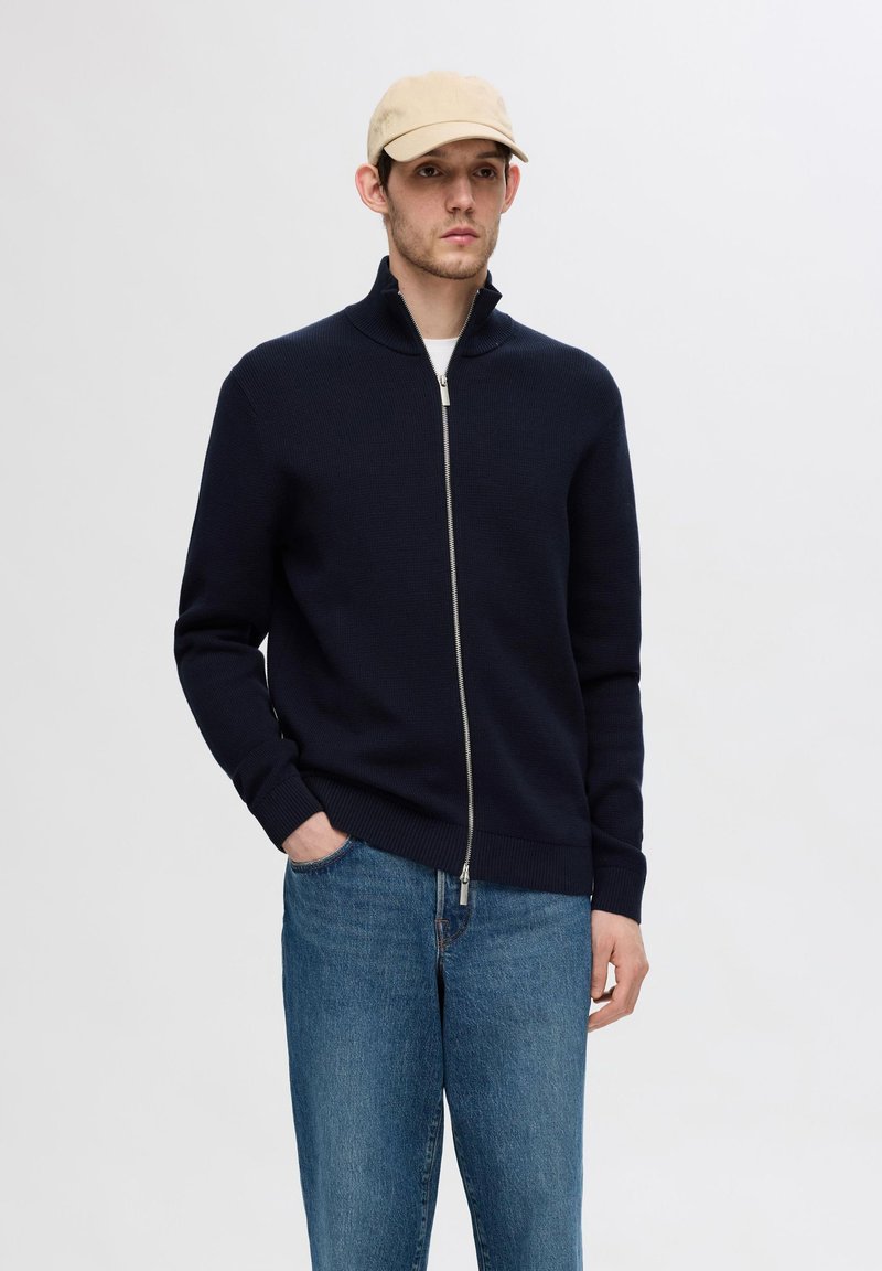 Maglione navy con zip, dalla texture a coste e colletto alto, abbinato a jeans di teak blu chiaro. Il modello indossa un cappello beige.