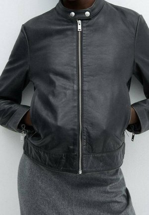 Osoba ubrana w czarną skórzaną kurtkę zapinaną na zamek błyskawiczny, z zatrzaskami przy kołnierzu i rękami wsuniętymi do bocznych kieszeni, w połączeniu z szarymi spodniami lub spódnicą.