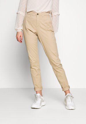 Femme portant un chino beige retroussé, des baskets blanches et un chemisier blanc à manches longues avec une texture à pois et des détails en dentelle.