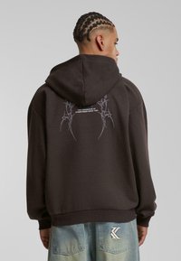 Sudadera marrón oscuro con un corte relajado, que presenta un bolsillo frontal tipo canguro y un diseño gráfico en la parte posterior en gris claro.
