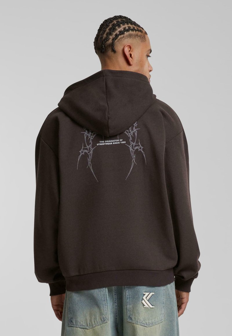 Sudadera marrón oscuro con un corte relajado, que presenta un bolsillo frontal tipo canguro y un diseño gráfico en la parte posterior en gris claro.