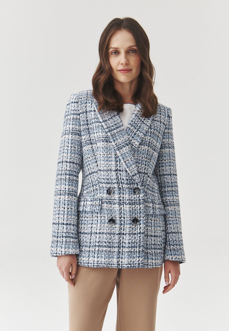 Mujer vestida con un blazer de cuadros azul y blanco de doble botonadura con botones negros y pantalones beige, de pie contra un fondo liso.