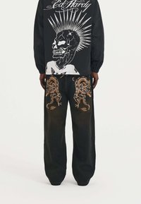 Camisa negra de manga larga con un gráfico de calavera blanca con rayos, combinada con pantalones de mezclilla oscuros adornados con un bordado detallado de dragón.