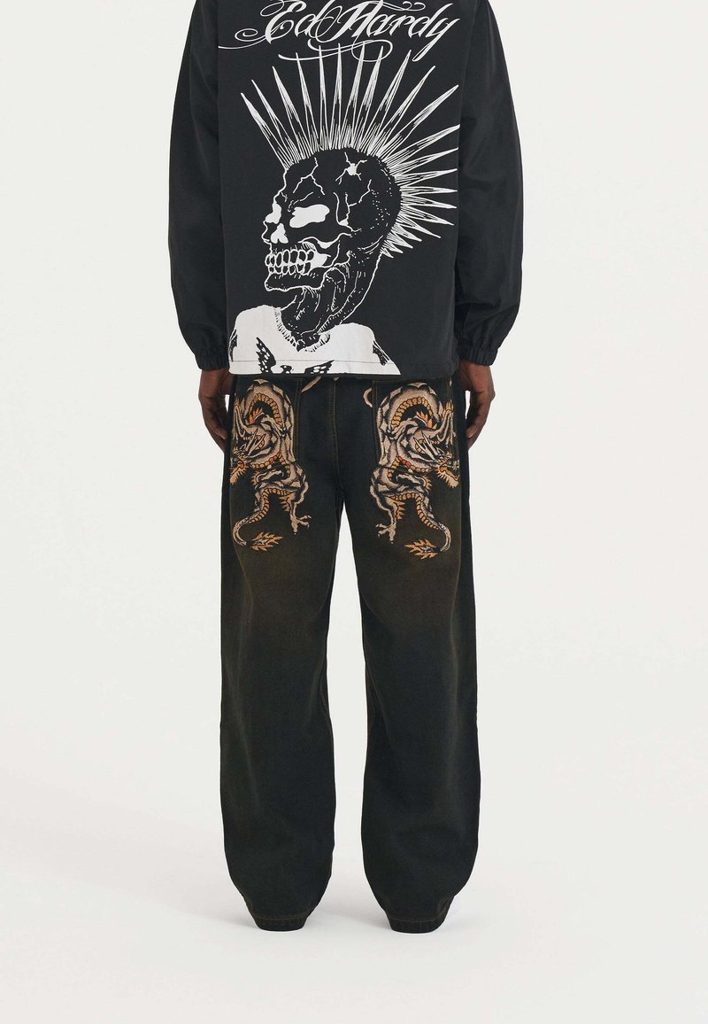 Camisa negra de manga larga con un gráfico de calavera blanca con rayos, combinada con pantalones de mezclilla oscuros adornados con un bordado detallado de dragón.