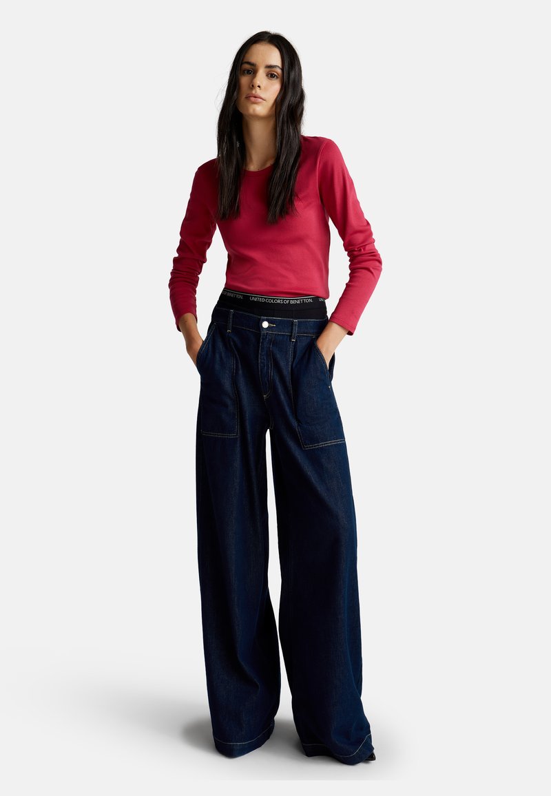 Donna con capelli lunghi e scuri, vestita con un top rosso a maniche lunghe e pantaloni in denim blu scuro a gamba larga, in piedi con le mani in tasca.