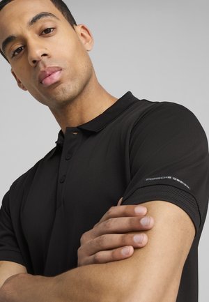 Mann mit verschränkten Armen, der ein schwarzes Polohemd mit kurzen Ärmeln von Porsche Design vor einem schlichten grauen Hintergrund trägt.