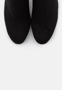 Un par de botas de tobillo de ante negro con punta afilada, vistas desde arriba sobre un fondo gris claro.