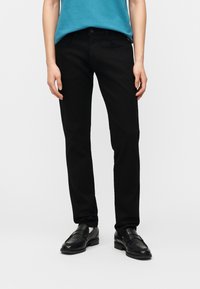 Personne portant un pantalon noir droit, des chaussures habillées noires et une chemise turquoise, debout devant un fond clair uni.