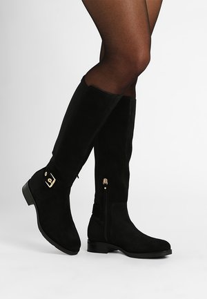 Jambes portant des collants noirs transparents et des bottes hautes en daim noir avec talons bas, boucle dorée et fermeture éclair latérale sur fond blanc.