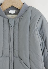Graue Kleinkindjacke mit gesteppter Textur, geripptem Kragen und Reißverschluss. Verfügt über zwei Fronttaschen und ein weiches Innenfutter.
