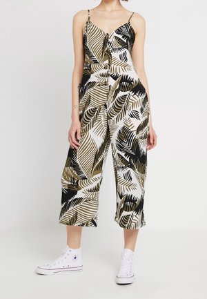 Woman wearing a sleeveless, wide-leg jumpsuit with black, white, and olive green tropical leaf pattern, paired with white high-top sneakers.