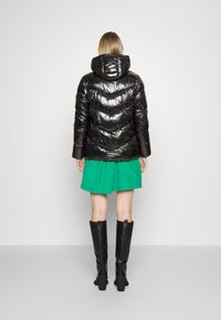 Chaqueta de plumas negra con una textura brillante y capucha, con un acolchado en forma de chevron. Combinada con una falda verde y botas negras hasta la rodilla.