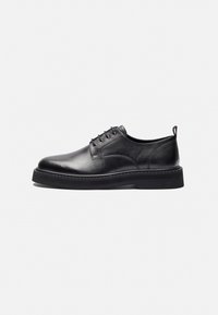 Zapato negro de cuero con cordones, punta redonda, detalles de costura y suela de goma gruesa. Presenta un perfil bajo y un discreto lazo en la parte trasera.