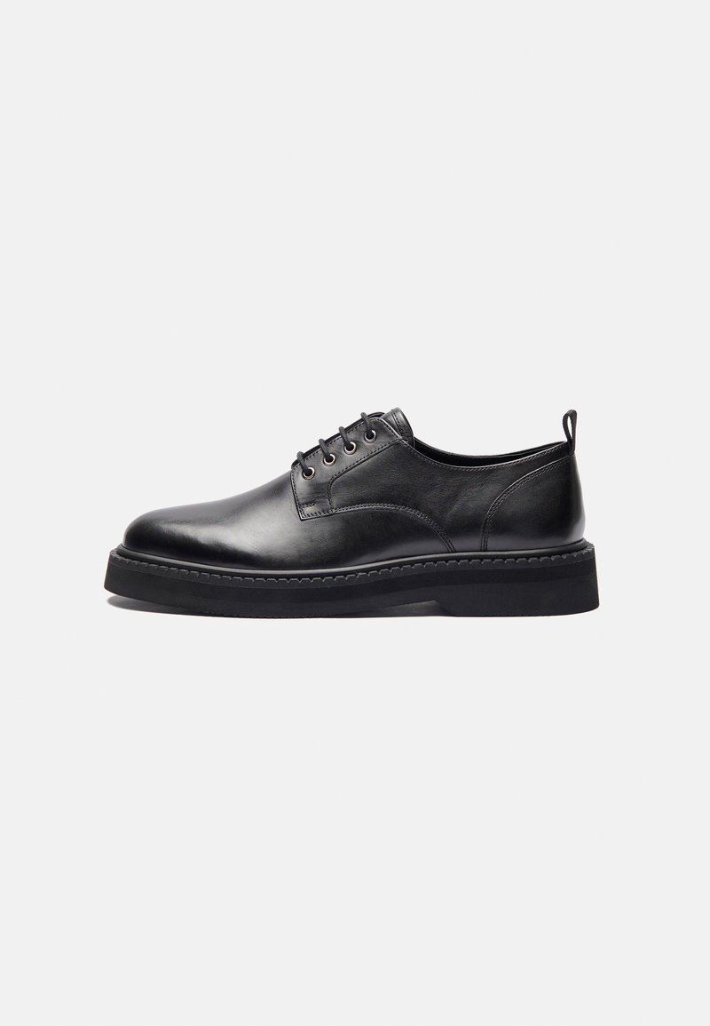 Zapato negro de cuero con cordones, punta redonda, detalles de costura y suela de goma gruesa. Presenta un perfil bajo y un discreto lazo en la parte trasera.