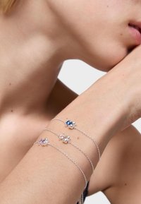 Tres delicadas pulseras de cadena de plata con pequeños charms de cuentas agrupadas en azul, púrpura y plata, llevadas en un antebrazo cerca de una cara.