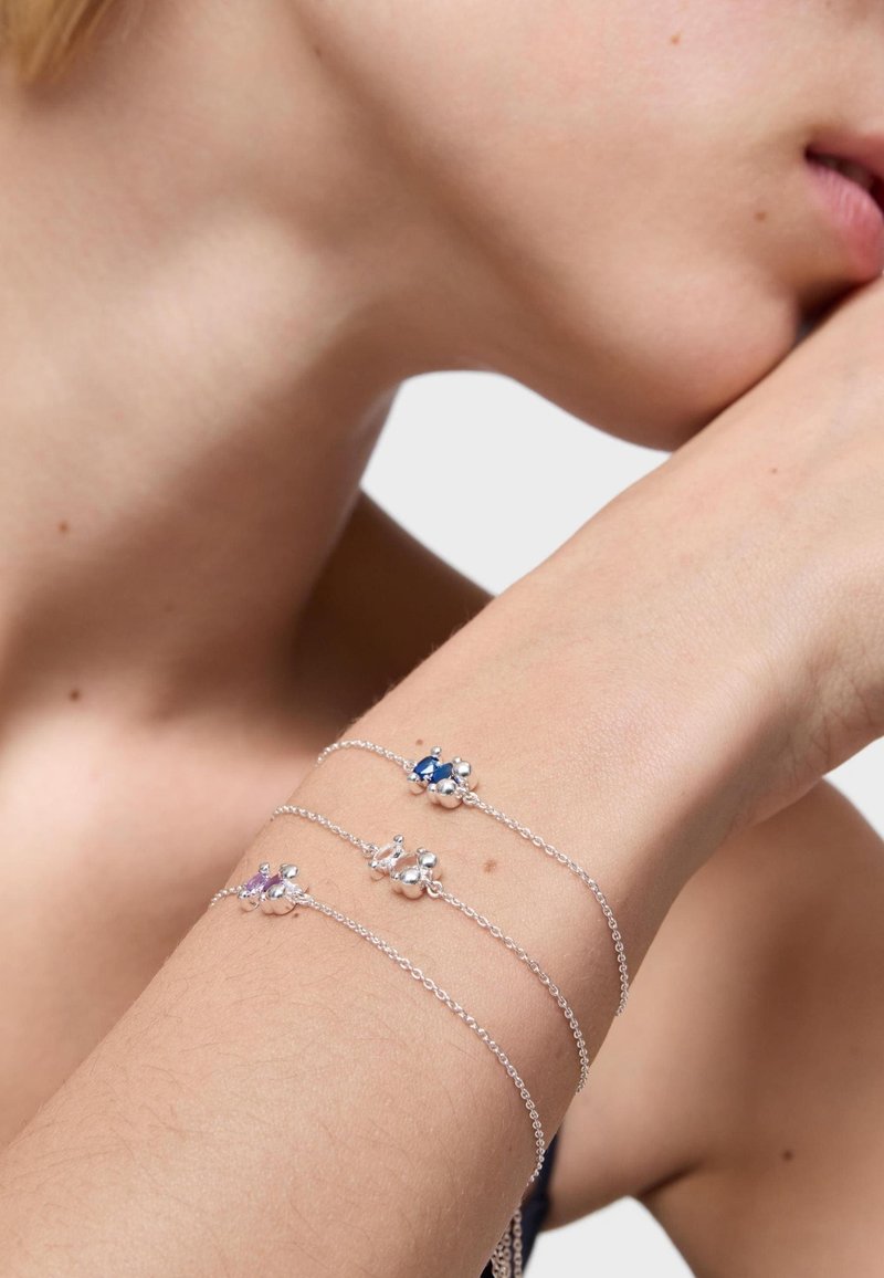 Tres delicadas pulseras de cadena de plata con pequeños charms de cuentas agrupadas en azul, púrpura y plata, llevadas en un antebrazo cerca de una cara.