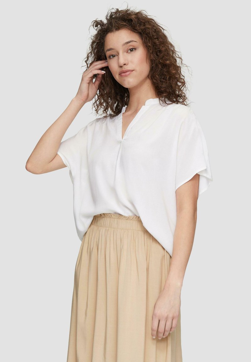 Blouse blanche à manches courtes avec un col en V et un tissu doux et fluide ; associée à une jupe beige plissée taille haute.