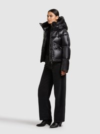 Zwarte pufferjas met een glanzende afwerking, getailleerd ontwerp, hoge kraag en zijzakken, gecombineerd met zwarte wijde jeans en laarzen met hak.