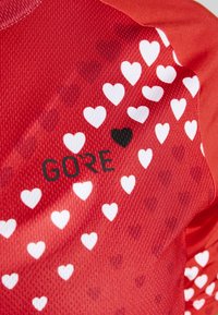 Czerwona teksturowana tkanina z rozrzuconymi białymi sercami, jednym czarnym sercem oraz czarnym napisem "GORE" drukowanym na powierzchni.