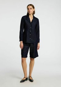 Camicia in denim scuro con bottoni e maniche lunghe abbinata a pantaloni corti fino al ginocchio. Tessuto liscio; leggera lucentezza; bottoni in metallo argentato.