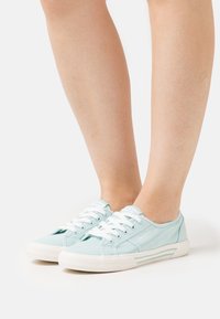 Ljusblå låga kanvas sneakers med vita snören och randiga sulor, bärs på barfotad, visad från sidan på en vit bakgrund.