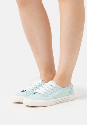 Helle blaue Canvas-Sneaker mit niedrigen Absätzen und weißen Schnürsenkeln sowie gestreiften Sohlen, die auf baren Füßen getragen werden, seitlich auf einem weißen Hintergrund gezeigt.