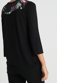 Musta pitkähihaine paita, jossa on kukkakuosiset yksityiskohdat selässä, pehmeä rakenne ja rento malli pyöristettyine helmoin.