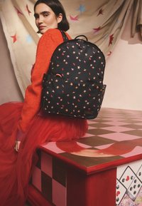 Moteris raudonu megztiniu ir tiulio sijonu sėdi ant dažyto šachmatų lentos stalo, vilkinti juodą kuprinę su raudonų, rožinių ir oranžinių žvaigždžių raštais.