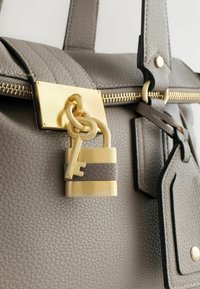 Harmaa teksturoitu nahkalaukku, jossa on kullanvärinen vetoketju ja lukonkoriste. Siinä on tasainen vetoketjun vedin, ketjusomiste ja riippuva tunnuslappu.